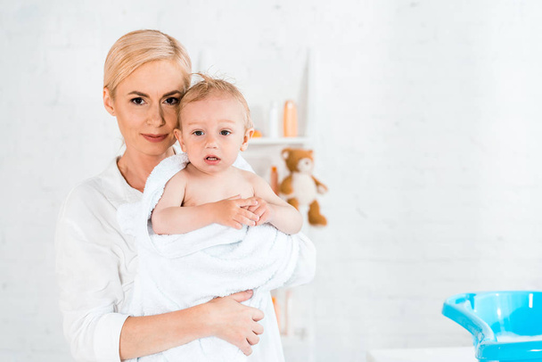 attractive blonde mother holding in arms cute toddler son in bathroom  - Zdjęcie, obraz