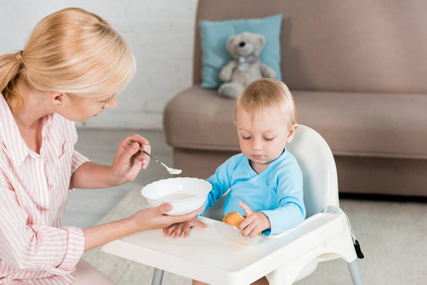 Blonde Mutter hält Schüssel und füttert Kleinkind-Sohn zu Hause  - Foto, Bild