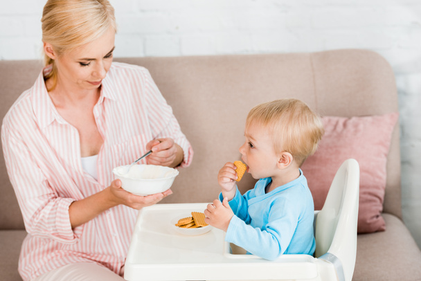 blonde mother holding spoon with baby food near cute toddler son sitting in feeding chair  - Фото, зображення