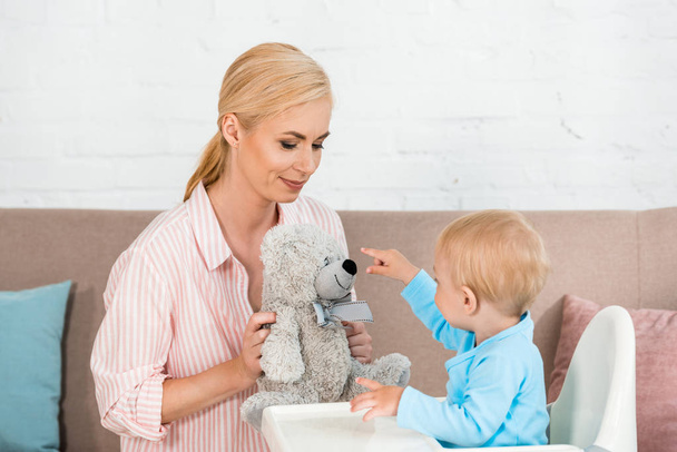 cute toddler son pointing with finger at teddy bear near cheerful mother - Foto, Bild