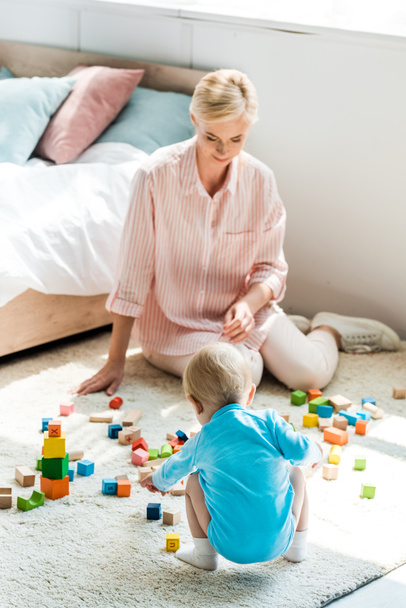happy mother sitting on carpet and looking at toddler son playing with building bricks  - Foto, Imagem