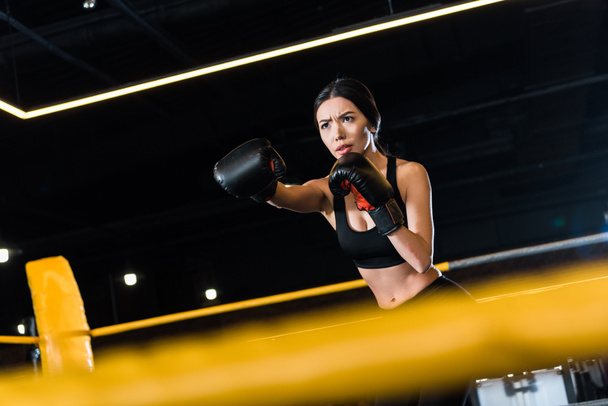 selective focus of serious woman boxing while standing in boxing gloves in gym  - Fotoğraf, Görsel