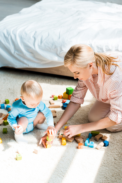 приваблива блондинка сидить на килимі біля дитячого сина, грає з будівельними цеглами
  - Фото, зображення