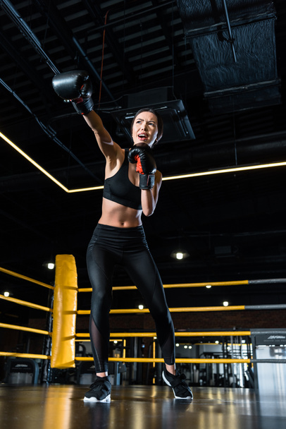 низкий угол зрения уверенной женщины бокс, стоя в боксёрских перчатках в тренажерном зале
  - Фото, изображение