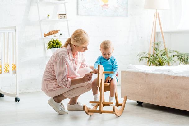 cute toddler kid on wooden rocking horse near attractive blonde mother  - Photo, Image