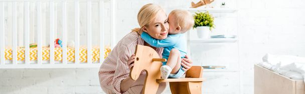panoramic shot of cute toddler son hugging happy mother near rocking horse  - Fotoğraf, Görsel