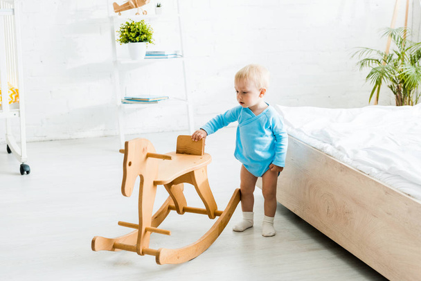cute toddler boy standing near wooden rocking horse in modern bedroom  - Photo, image