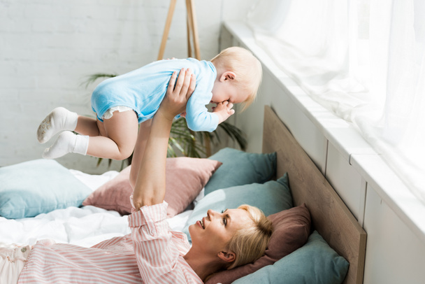 happy mother holding in arms cute toddler son while lying on bed  - Fotó, kép
