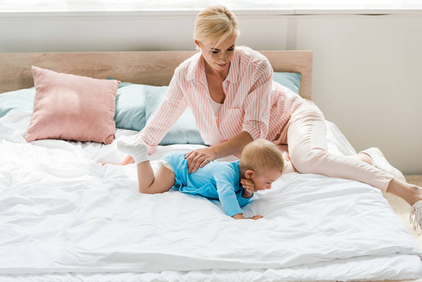 Veselá blondýnka dívaje se na roztomilého syna plazících se po posteli  - Fotografie, Obrázek