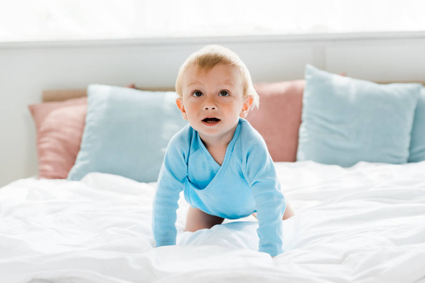 χαριτωμένο νήπιο παιδί σέρνεται στο κρεβάτι με λευκά κλινοσκεπάσματα και μαξιλάρια στο σπίτι  - Φωτογραφία, εικόνα