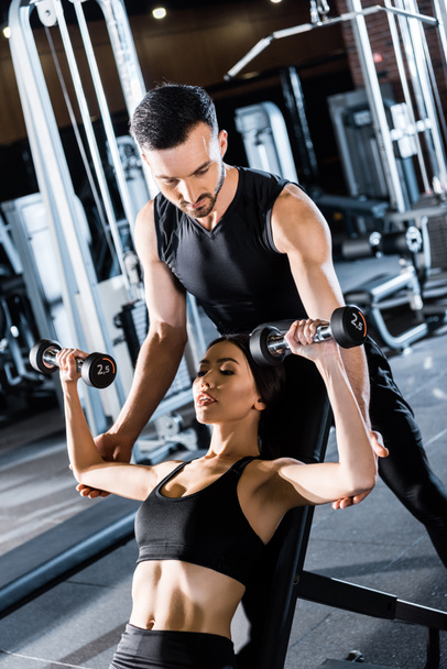 selective focus of young athletic woman working out with dumbbells near handsome  trainer in gym  - Foto, Bild