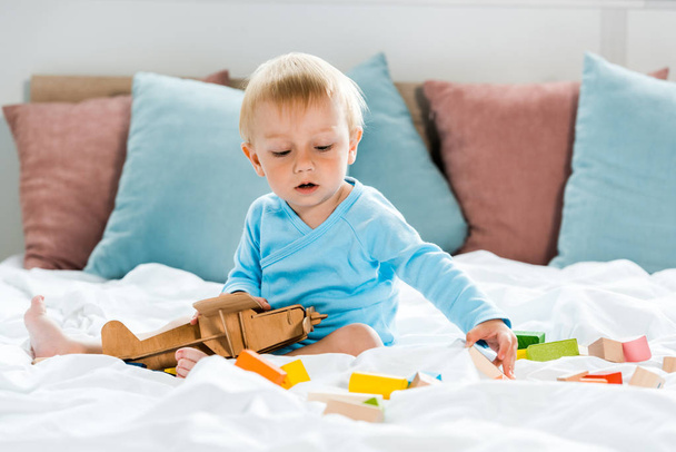 νήπιο παιδί που παίζει με ξύλινο διαεροπλάνο κοντά σε πολύχρωμα μπλοκ παιχνιδιών στο κρεβάτι  - Φωτογραφία, εικόνα