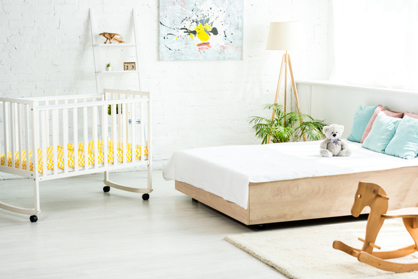 Babybett neben Bett mit weißer Bettwäsche und Kissen in der Nähe von Teddybär und Schaukelpferd  - Foto, Bild