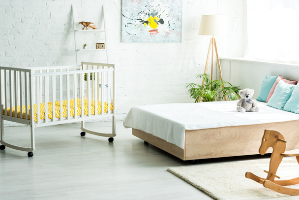 baby crib near bed with white bedding and pillows near teddy bear and wooden rocking horse  - Photo, Image