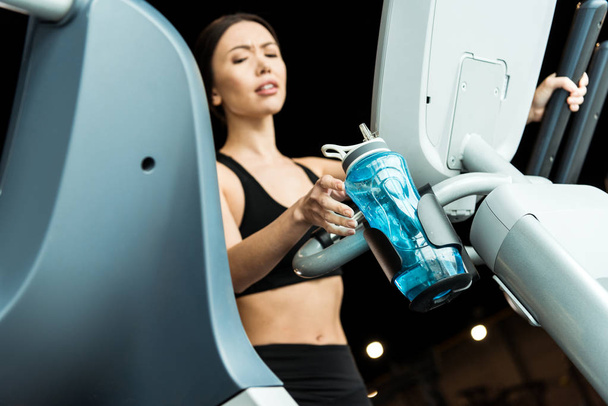 selective focus of athletic woman taking sport bottle while working out on exercise bike  - Photo, Image