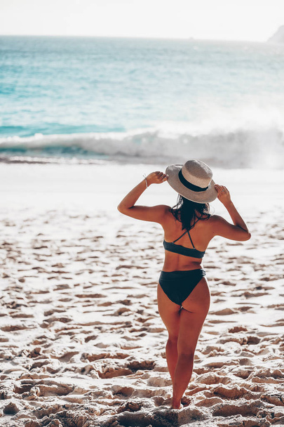 девушка в черном бикини стоит на белоснежном пляже двумя руками, держа на голове белую шляпу - Фото, изображение