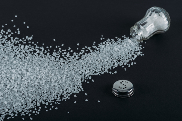 top view of sprinkled salt crystals near glass salt shaker on black background - Photo, Image