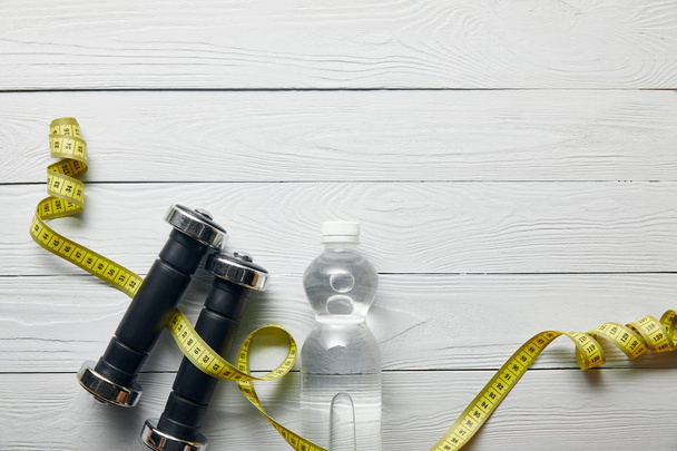 top view of bottle with water, yellow measuring tape and dumbbells on wooden white background with copy space - Foto, Imagen