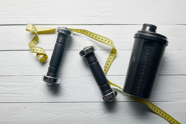 top view of black shaker cup, yellow measuring tape and dumbbells on wooden white background  - Foto, afbeelding