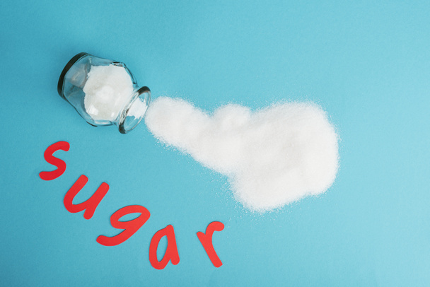 Top weergave van rood papier gesneden woord suiker in de buurt van glazen pot en bespelde suiker kristallen op blauwe achtergrond - Foto, afbeelding