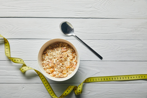 top view of measuring tape, breakfast cereal in bowl near spoon on wooden white background with copy space - Φωτογραφία, εικόνα