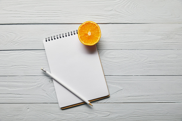 top view of orange half, blank notebook and pencil on wooden white background with copy space - Photo, Image