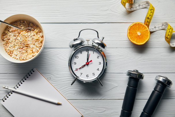 vue du dessus du réveil argenté, carnet avec crayon, céréales pour petit déjeuner, ruban à mesurer, haltères et moitié orange sur fond blanc en bois
  - Photo, image