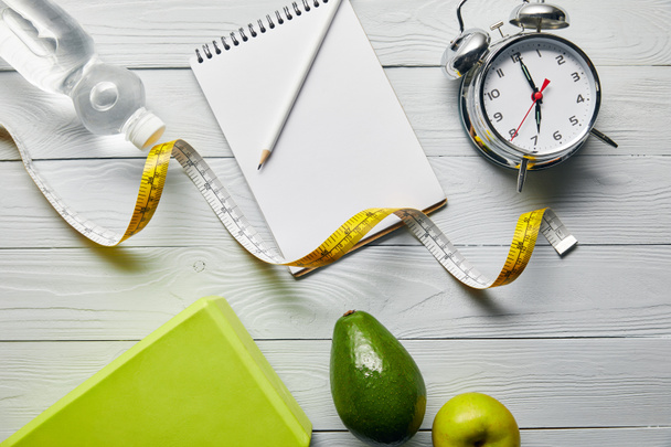 bovenaanzicht van notebook en potlood, alarm klok, meetlint, avocado, appel en water op houten witte achtergrond  - Foto, afbeelding