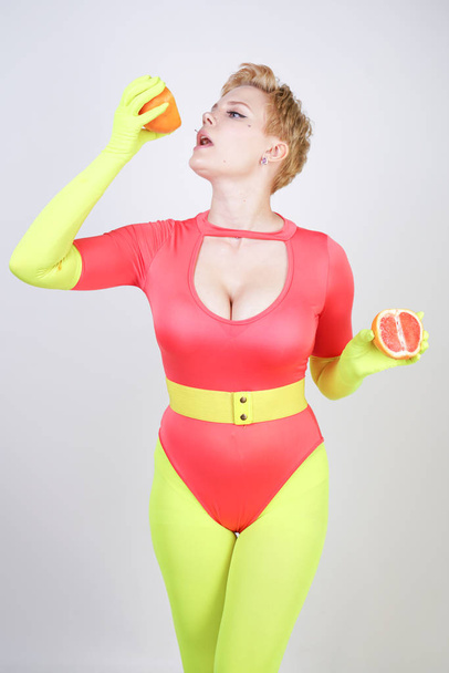 alternative caucasian girl with short hair dressed in sporty red spandex bodysuit and bright tights with green neon gloves. curvaceous woman holds a sliced grapefruit on a white studio background - Photo, Image