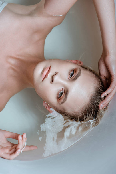 top view of sexy woman looking at camera while lying in clear water in bathtub - Фото, изображение