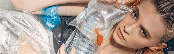 panoramic shot of sad wet woman holding goldfish in plastic bag among rubbish in bathtub - Фото, зображення