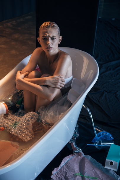 sad wet woman sitting in bathtub with trash and looking at camera - Photo, Image