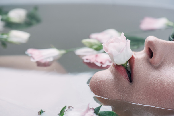 partial view of beautiful woman lying in clear water with rose in mouth - Foto, Imagen