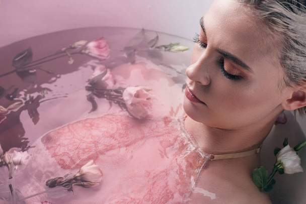 beautiful woman lying in clear water with flowers in white bathtub, pink toned image - Фото, зображення