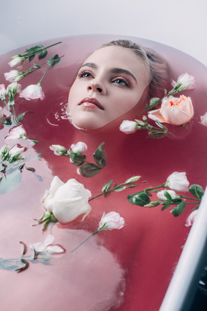 kaunis nuori nainen makaa kylpyammeessa värillisellä vaaleanpunaisella vedellä ja kukilla ja katsoo pois
 - Valokuva, kuva