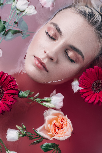 top view of beautiful young woman with closed eyes lying in bathtub with colored pink water and flowers - Photo, Image