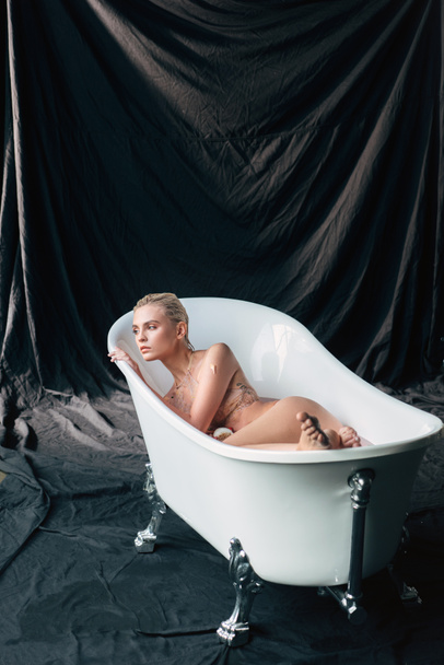 beautiful wet and sexy blonde woman lying in bathtub and looking away on black background - Photo, Image