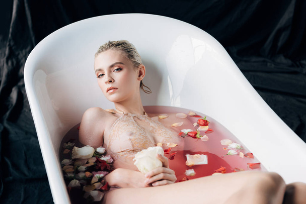 beautiful wet and sexy blonde woman lying in bathtub with pink water and petals  - Fotografie, Obrázek