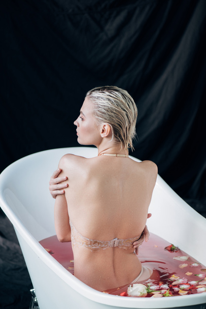 back view of blonde sexy wet woman sitting in bath with pink water and rose petals - Photo, Image
