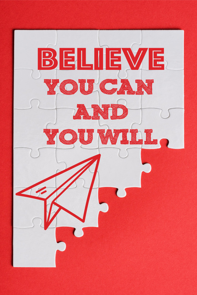 top view of white connected puzzle pieces with paper plane and believe you can and you will lettering isolated on red  - Photo, Image