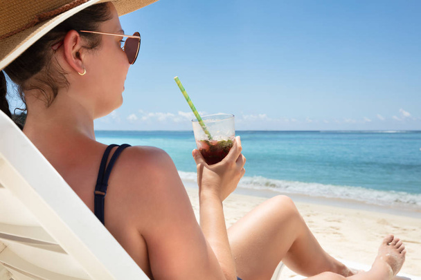 Young Woman In Hat Sitting On White Deck Chair Enjoying The Juice Drink On Beach - Foto, Imagem