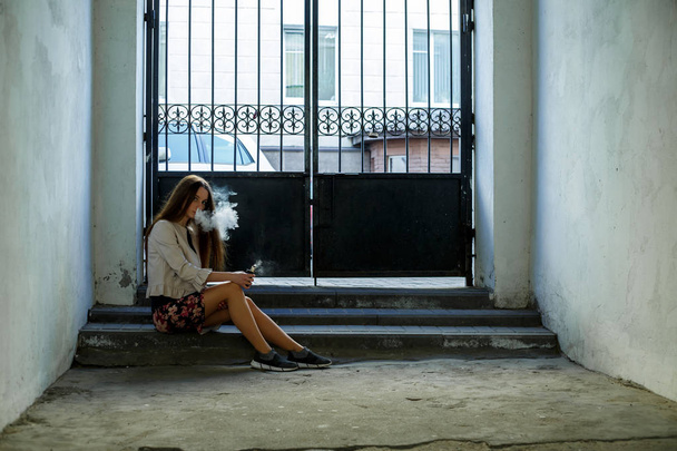 Vape teenager. Young cute girl in casual clothes smokes an electronic cigarette near the wall in prison cell. Bad habit that is harmful to health. Vaping activity. - Photo, image