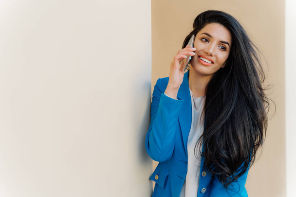 Happy female model with dark long hair calls via cell phone and talks in roaming wears blue jacket looks aside has thoughtful expression. Office worker speaks on cell phone. Business and communication - Фото, зображення