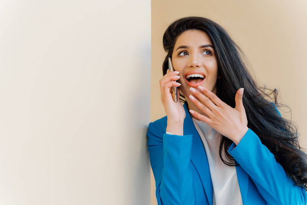 Positive business lady open mouth from interest, touches chin, looks aside, fascinated by rumors, has telephone conversation with close friend, gossip about latest news, wears formal blue costume - Fotoğraf, Görsel