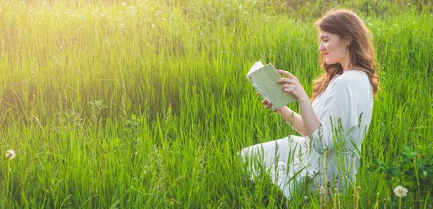 Το όμορφο κορίτσι στο πεδίο διαβάζει ένα βιβλίο. Το κορίτσι που καθόταν σε ένα γρασίδι, διαβάζοντας ένα βιβλίο. Ανάπαυση και ανάγνωση - Φωτογραφία, εικόνα