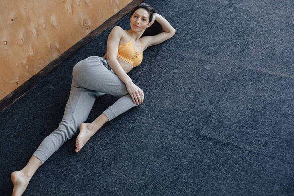 Young attractive fitness girl lying on the floor near the window on the background of a wooden wall, resting on yoga classes - Fotografie, Obrázek