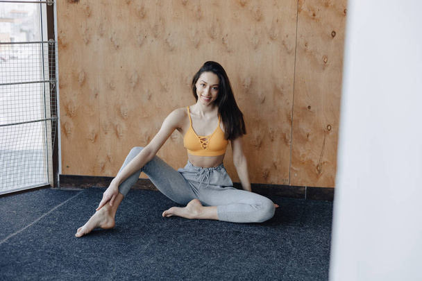 Young attractive fitness girl sitting on the floor near the window on the background of a wooden wall, resting on yoga classes - Foto, afbeelding