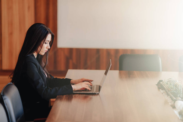 Jonge aantrekkelijke emotionele meisje in Business-stijl kleren zitten aan een bureau op een laptop en telefoon in het kantoor of Auditorium - Foto, afbeelding