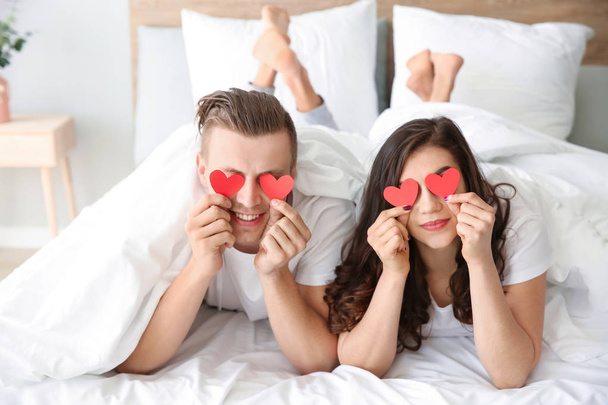 Ευτυχισμένο νεαρό ζευγάρι με χάρτινες καρδιές ξαπλωμένοι στο κρεβάτι - Φωτογραφία, εικόνα