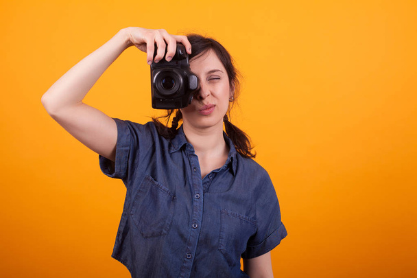 Attractive caucasian woman with photo camera focusing in studio over yellow background - Photo, Image